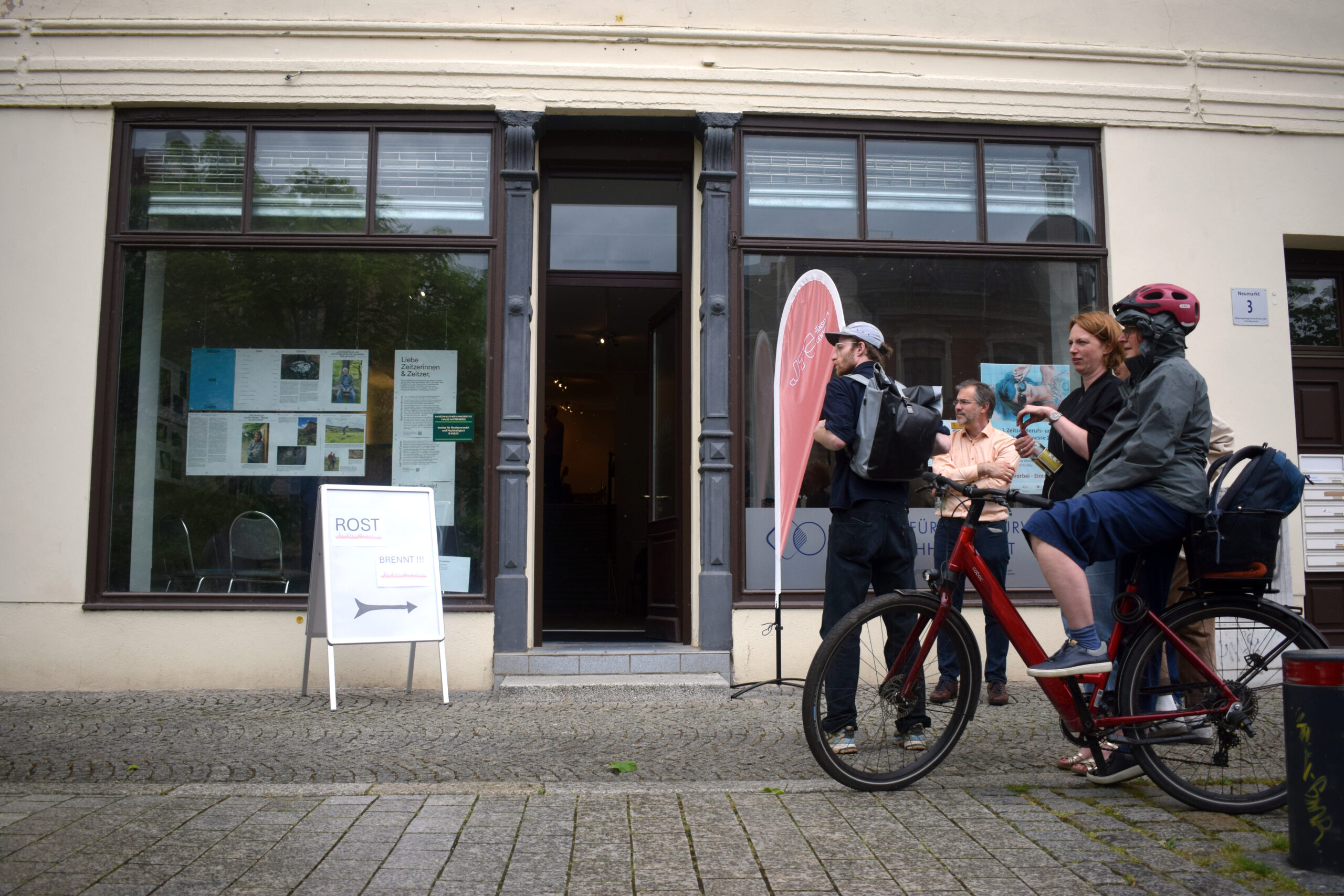 Menschen stehen vor einem Ladenlokal mit großen Schaufenstern und offener Tür. Eine Person hat ein Fahrrad dabei.